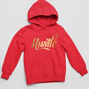 Puede incluir: Una sudadera con capucha de color rojo brillante que presenta mangas largas, un bolsillo tipo canguro frontal y el texto "I AM THE Hustle" impreso en una fuente dorada metálica en el centro del pecho. La prenda tiene capucha y puños acanalados.