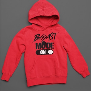 Puede incluir: Una sudadera con capucha de color rojo brillante y manga larga que incluye un bolsillo tipo canguro. En el pecho presenta el texto en negro BEAST MODE ON, con la palabra ON dentro de un gráfico negro en forma de píldora junto a un círculo blanco.