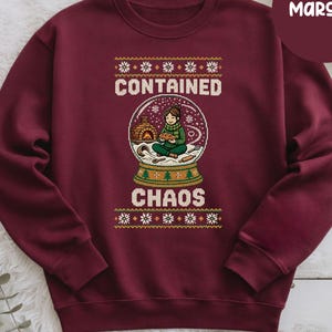May include: A maroon crewneck sweatshirt featuring a graphic of a snow globe containing a person baking bread in front of an oven. The text "CONTAINED CHAOS" is printed above and below the globe, surrounded by white snowflake patterns.