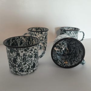 May include: Three enamel mugs featuring a black and white speckled marble pattern. Each mug has a cylindrical body, a curved handle on the side, and a solid black rim. The mugs are arranged with two standing upright and one lying on its side.
