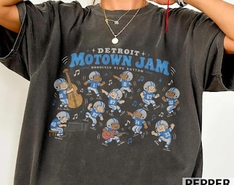 May include: A charcoal gray t-shirt featuring a graphic print with the text "DETROIT MOTOWN JAM" and "HONOLULU BLUE RHYTHM". The design includes multiple cartoon figures in blue football uniforms playing musical instruments and dancing.