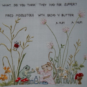 May include: Embroidered artwork depicting a whimsical scene with a frog, a mouse, and a ladybug. The artwork features a light blue background with green, pink, and red embroidery. The text "AND WHAT DO YOU THINK THEY HAD FOR SUPPER? FRIED MOSQUITOES WITH BREAD 'N' BUTTER HUM A HUM A HUM TAETIA" is embroidered in black thread.