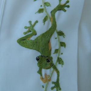 May include: Green embroidered frog hanging upside down on a green vine with leaves. The frog has black eyes and a yellow belly.