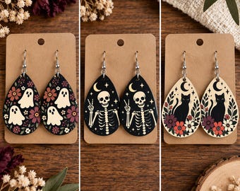 May include: Three pairs of teardrop-shaped dangle earrings on brown display cards. The first pair features white ghosts and flowers on a black background. The second pair shows a skeleton making a peace sign with stars and a moon. The third pair displays a black cat with flowers and a crescent moon.