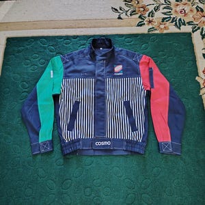 May include: A navy blue jacket featuring a vertical white pinstripe pattern on the front, a green right sleeve, and a red left sleeve. The garment includes a stand-up collar, two front pockets, and the word "cosmo" printed on the chest and hem.