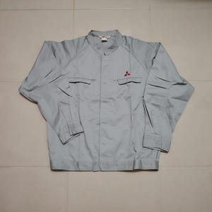May include: A light gray long-sleeve work jacket featuring a mandarin collar, a concealed front button placket, and two chest pockets with flaps. A small red three-diamond logo is embroidered on the upper left chest area of the garment.