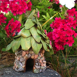 May include: A charming fairy house sculpture featuring a leaf-shaped roof and a stone base. The green roof is decorated with pink flowers and a ladybug. The house is set amidst vibrant red flowers and foliage.
