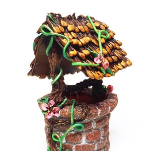 May include: A miniature wishing well made of clay with a brown brick exterior, a thatched roof, and green vines with pink flowers.