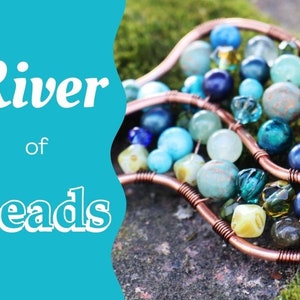 May include: A copper wire sculpture with a river of blue, green, and yellow beads. The sculpture is shaped like two curved lines, and the beads are arranged in a flowing pattern. The sculpture is sitting on a bed of green moss.