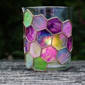 May include: A glass candle holder with a hexagonal pattern of pink, purple, green, and blue paint. The candle holder is on a wooden surface.