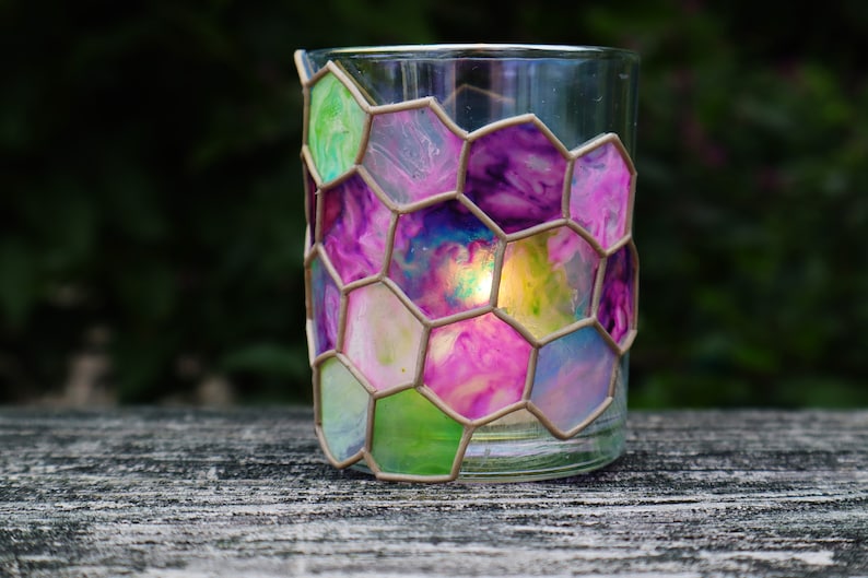 May include: A glass candle holder with a hexagonal pattern of pink, purple, green, and blue paint. The candle holder is on a wooden surface.