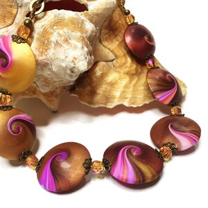 May include: A handcrafted necklace featuring large, oval and round beads in shades of brown, gold, and pink. Each bead has a swirling design. The necklace is accented with smaller, amber-colored beads and antique brass-toned metal findings.