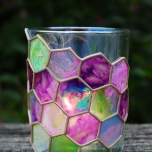 May include: A glass candle holder with a hexagonal pattern. The glass is painted with shades of pink, purple, green, and blue. The candle holder is sitting on a wooden surface.