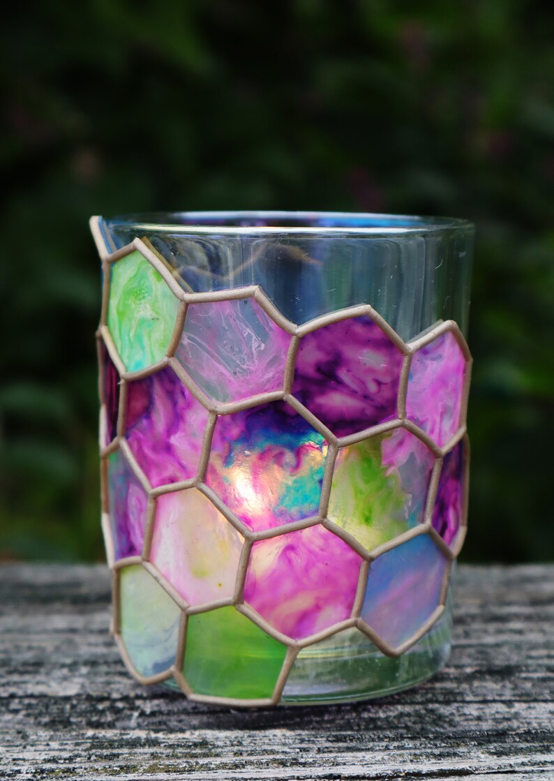 May include: A glass candle holder with a hexagonal pattern. The glass is painted with shades of pink, purple, green, and blue. The candle holder is sitting on a wooden surface.