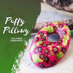 May include: A pink polymer clay pendant with a floral design. The pendant has a hole in the centre and is shaped like a doughnut. The flowers are red with black centres and green leaves.