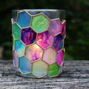 May include: A glass candle holder with a honeycomb design. The glass is painted in shades of blue, green, pink, and purple. A candle is burning inside the holder.