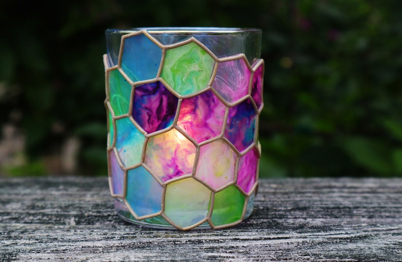 May include: A glass candle holder with a honeycomb design. The glass is painted in shades of blue, green, pink, and purple. A candle is burning inside the holder.