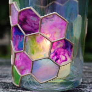 May include: A glass candle holder with a honeycomb design. The glass is painted with shades of pink, blue, and green. A candle is burning inside the holder.