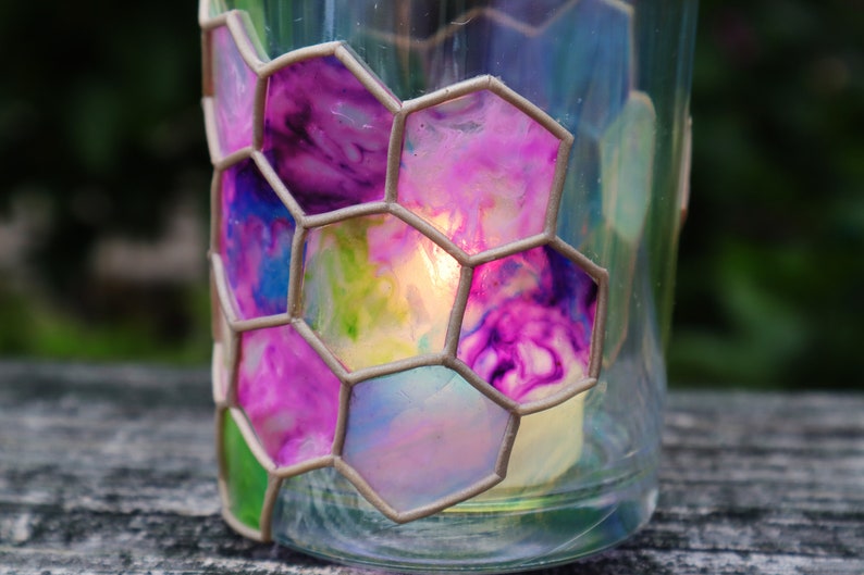 May include: A glass candle holder with a honeycomb design. The glass is painted with shades of pink, blue, and green. A candle is burning inside the holder.