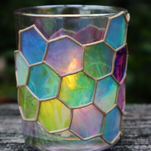 May include: A glass candle holder with a hexagonal pattern. The glass is painted with shades of blue, green, yellow, and pink. The candle holder is lit and the light shines through the glass.