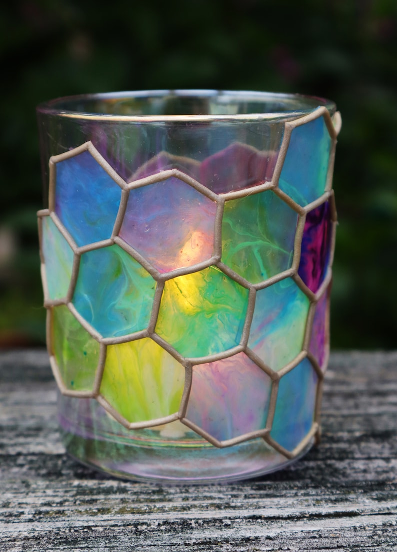 May include: A glass candle holder with a hexagonal pattern. The glass is painted with shades of blue, green, yellow, and pink. The candle holder is lit and the light shines through the glass.