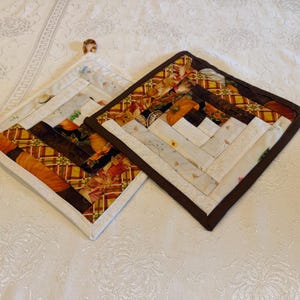 May include: Two square quilted pot holders featuring a log cabin pattern. The fabric displays autumn-themed prints including pumpkins, leaves, and plaid in shades of orange, brown, cream, and yellow. Each pot holder has a small fabric hanging loop.