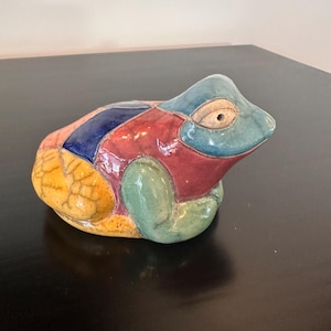 May include: A ceramic frog figurine featuring a patchwork design with glossy sections in blue, red, green, and yellow. The yellow portion displays a dark crackle glaze pattern, while the head is a solid blue with a beige eye area.