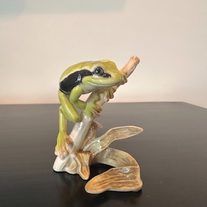 May include: A ceramic figurine of a green frog with a prominent black stripe along its side, perched on a light brown textured branch. The frog has a smooth, glossy finish and is depicted in a resting position with its limbs gripping the branch.