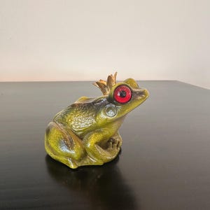 May include: A green frog figurine with a textured skin surface, large prominent red eyes, and a small gold-colored crown positioned on the top of its head. The frog is depicted in a seated position with its legs tucked underneath its body.