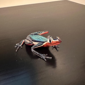 May include: A glass frog figurine featuring a metallic teal back, a reddish-orange head, and dark blue limbs. The sculpture has large, prominent eyes with black pupils and clear glass feet, showcasing a smooth, reflective surface throughout the body.