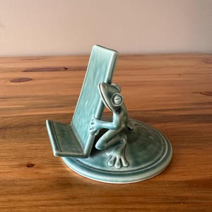 May include: A teal ceramic phone stand featuring a sculpted frog figure attached to a circular base. The frog is positioned as if holding up the rectangular support ledge, which is designed to hold a mobile device in an upright position.