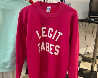 May include: Pink sweatshirt with the text "LEGIT BABES" in white lettering. The sweatshirt has a small red tag with the letter "R" on the bottom right corner.