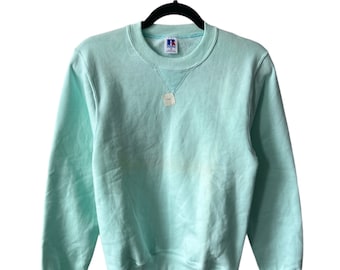 May include: A light blue crew neck sweatshirt with a Russell Athletic logo on the front. The sweatshirt has long sleeves and a ribbed hem and cuffs.