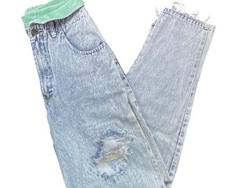 May include: Light blue acid wash denim jeans with a distressed knee and a green waistband. The jeans have a small embroidered detail near the pocket. The jeans are folded and displayed on a white background.