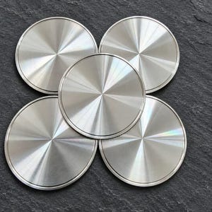 May include: A set of five circular coasters made of brushed stainless steel. Each coaster features a radial, sunburst-like texture that reflects light from the center outward, creating a metallic sheen across the flat, round surfaces.