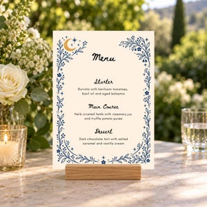 May include: A cream-colored menu card held by a wooden stand, featuring a blue floral border with stars and a crescent moon. The text reads Menu, Starter: Burrata with heirloom tomatoes, basil oil and aged balsamic, Main Course: Herb-crusted lamb with rosemary jus and truffle potato puree, Dessert: Dark chocolate tart with salted caramel and vanilla cream.