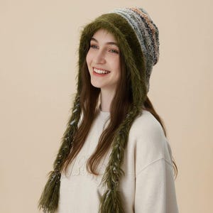May include: A knit beanie with long, fuzzy olive green earflaps and a multi-colored textured crown featuring shades of grey, blue, and tan. The hat has a soft, shaggy texture around the face and hangs down with two long, braided-style side panels.