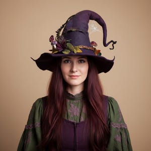 May include: A dark purple felted hat with a wide, curved brim and a pointed, drooping top. The hat is decorated with small fabric mushrooms, green leaves, and a white spiderweb embroidery detail on the side of the crown.