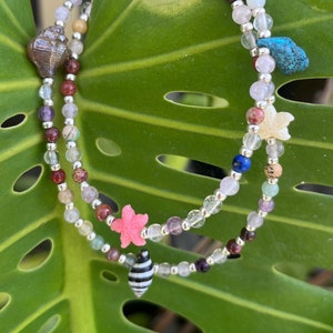Può includere: Due collane di perline con ciondoli a tema marino, tra cui conchiglie e stelle marine. Le collane presentano una varietà di perline colorate e sono esposte su una grande foglia verde. I ciondoli includono una conchiglia marrone, una stella marina rosa e una conchiglia a strisce bianche e nere.