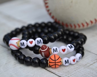 May include: Two black beaded bracelets with white letter beads spelling "MOM" and a heart charm. Each bracelet has a different sports charm: a baseball, a football, and a basketball.