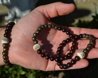May include: Two beaded bracelets, one brown with gold accents and one dark brown, with white beads. The bracelets are on a person's hand.