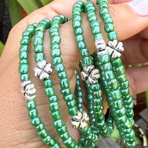 Könnte beinhalten: Mehrere grüne Perlenarmbänder mit silbernen Kleeblatt-Anhängern. Die Armbänder bestehen aus kleinen, runden, irisierenden grünen Perlen. Jedes Armband hat einen silbernen Kleeblatt-Anhänger.