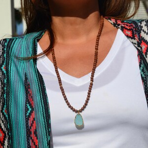 May include: A brown wooden beaded necklace with a turquoise pendant. The necklace is worn by a person wearing a white shirt and a turquoise and black patterned kimono.