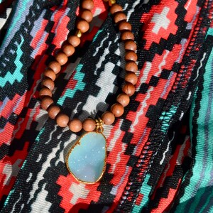 May include: A wooden beaded necklace with a light blue gemstone pendant. The necklace is on a colorful patterned fabric.
