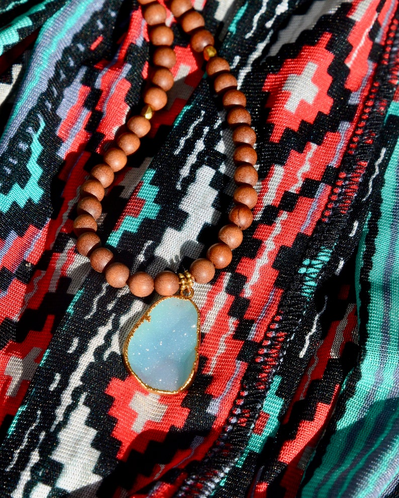 May include: A wooden beaded necklace with a light blue gemstone pendant. The necklace is on a colorful patterned fabric.
