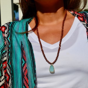 May include: A brown wooden beaded necklace with a light blue gemstone pendant. The necklace is worn by a person wearing a white shirt and a turquoise and black patterned cardigan.