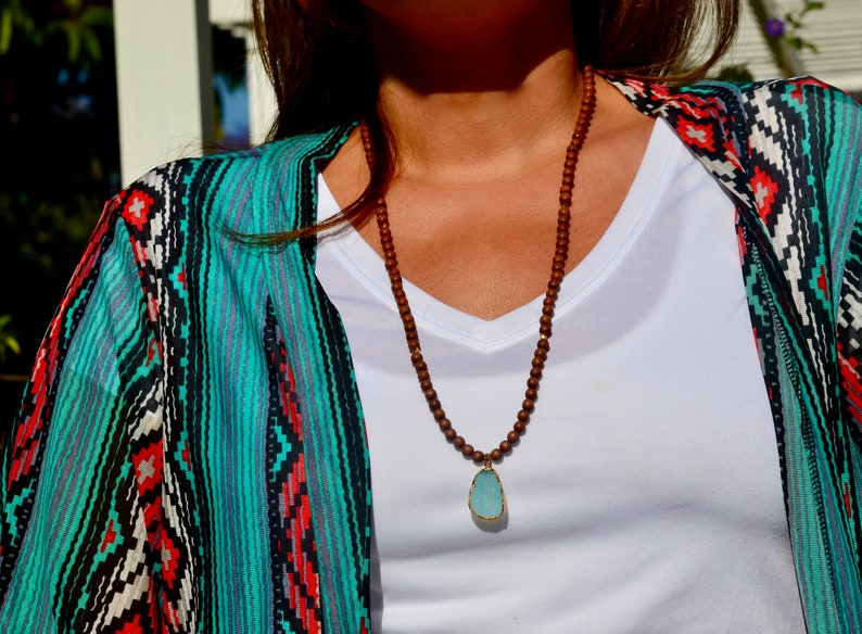 May include: A brown wooden beaded necklace with a light blue gemstone pendant. The necklace is worn by a person wearing a white shirt and a turquoise and black patterned cardigan.