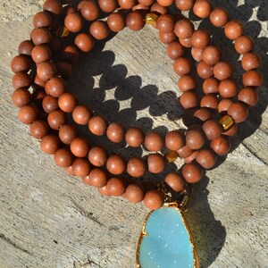 May include: A brown wooden beaded necklace with a light blue gemstone pendant. The pendant is set in gold and has a sparkly finish.