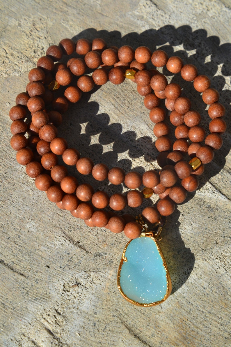 May include: A brown wooden beaded necklace with a light blue gemstone pendant. The pendant is set in gold and has a sparkly finish.