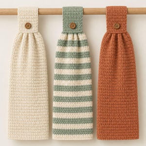 Op de afbeelding: Drie hangende gehaakte keukendoeken met een textuursteekpatroon. Eén doek is effen crème, één heeft horizontale strepen in saliegroen en crème, en één is effen terracotta. Elke doek heeft een lus aan de bovenkant die is vastgezet met een bruine knoop.